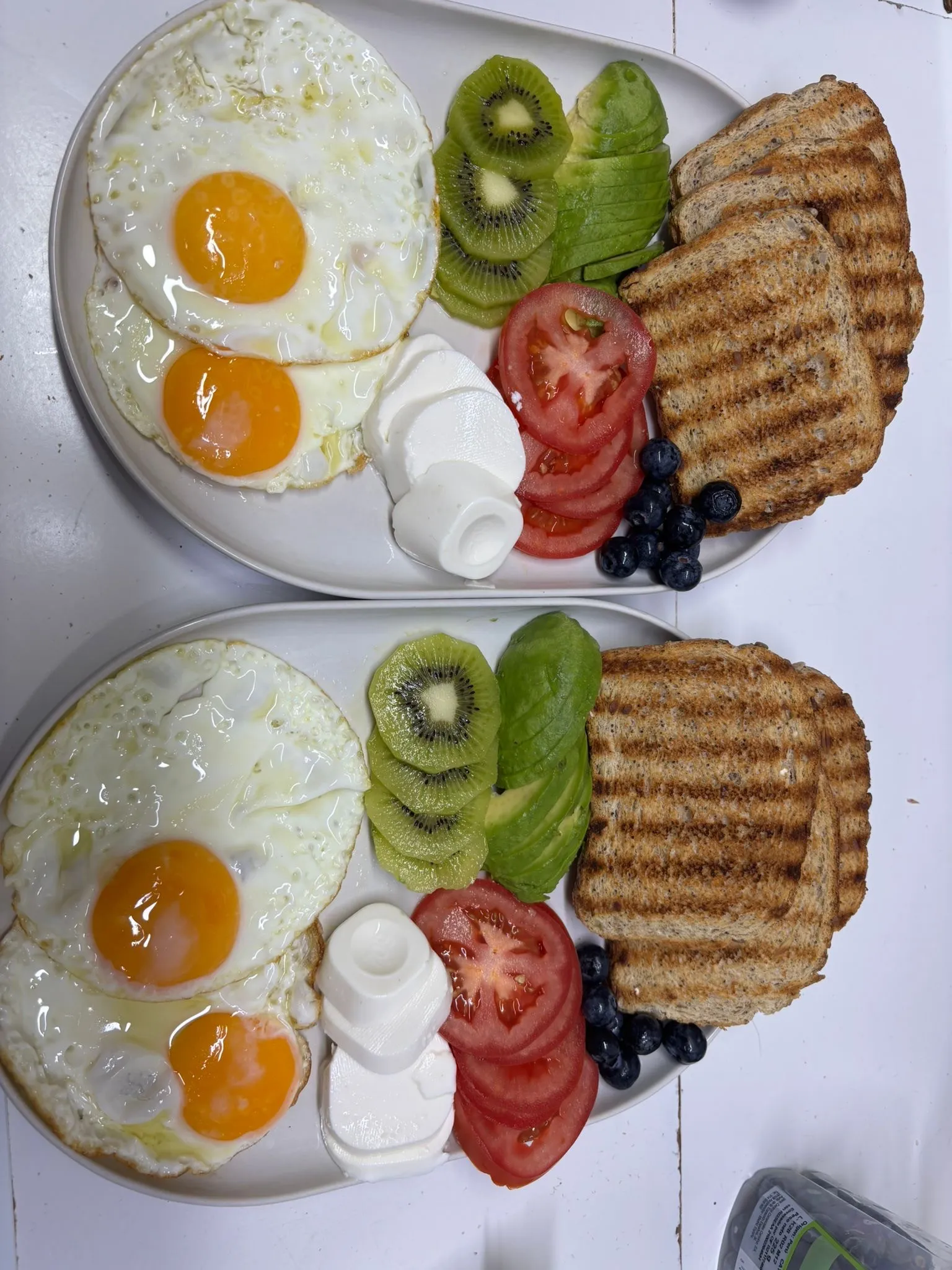 Dos desayunos con huevos fritos, tostadas, kiwi, aguacate, tomate y arándanos